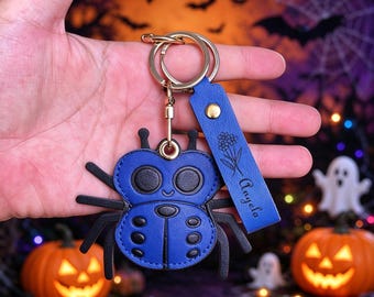 May include: A blue and black faux leather spider keyring featuring a circular metal ring and a matching blue rectangular strap. The strap is embossed with a floral design and the name Angelo in black script, secured by a gold-toned metal stud.