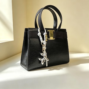 May include: A black structured handbag with a textured lizard-skin pattern, featuring two top handles and a gold-tone metal clasp. A white rope charm with black speckles and a silver-toned metallic balloon dog pendant hangs from one handle.
