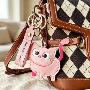 Könnte beinhalten: Ein rosafarbener katzenförmiger Schlüsselanhänger mit passendem rosa Riemen, der den Text Angela-Gugu trägt, befestigt an einer braun-cremefarbenen Tasche mit Argyle-Muster, braunem Lederbesatz und goldfarbenen Metallbeschlägen, inklusive Karabiner und Ringen.