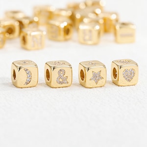 May include: A collection of gold-toned cube beads featuring various engraved symbols including a crescent moon, an ampersand, a star, and a heart, each inlaid with small clear crystals. The beads are arranged in a row with additional beads blurred in the background.