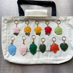 May include: Eleven assorted flower and leaf shaped keychains with gold-toned metal rings arranged on a cream-colored canvas tote bag. The keychains feature various colors including pink, yellow, red, green, blue, and orange, each with a small green stem.