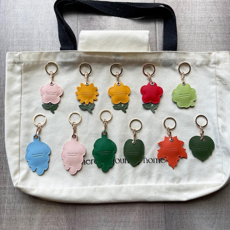 May include: Eleven assorted flower and leaf shaped keychains with gold-toned metal rings arranged on a cream-colored canvas tote bag. The keychains feature various colors including pink, yellow, red, green, blue, and orange, each with a small green stem.