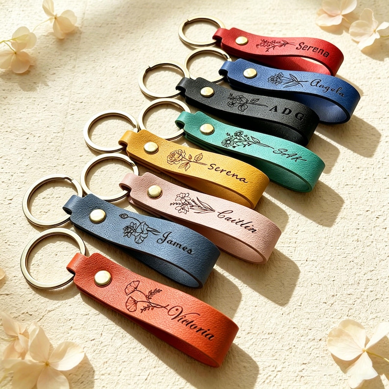 May include: A collection of seven leather keychains in red, blue, black, teal, yellow, pink, and orange. Each keychain features a gold-tone metal ring, a gold-tone snap button, and a custom engraved floral design with a name or initials.