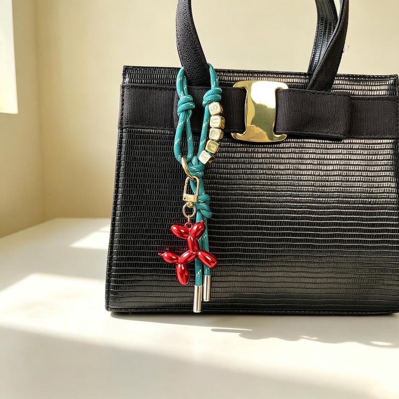 May include: A black textured handbag with a gold-tone metal clasp and a black fabric bow. Attached to the handle is a teal rope charm featuring gold-tone letter beads spelling SURF and a glossy red balloon dog pendant with a gold-tone clip.