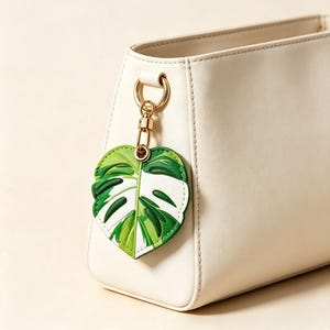 Puede incluir: Un bolso de color crema con un anillo de metal dorado y un cierre de mosquetón. Del cierre cuelga un dije en forma de corazón diseñado para parecerse a una hoja de monstera variegada con tonos de verde oscuro, verde claro y blanco.