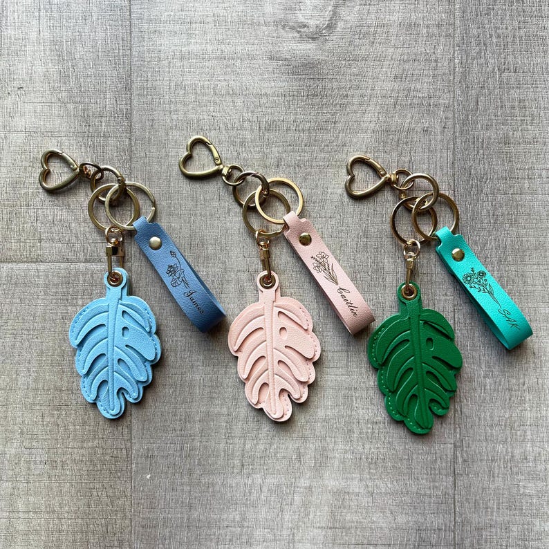 May include: Three keychains featuring heart-shaped gold-tone rings and leaf-shaped charms in light blue, pale pink, and green. Each keychain includes a matching leather-like strap engraved with a floral design and a name: James, Caitlin, and SAR.