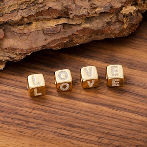May include: Four gold-toned cube-shaped beads arranged to spell the word LOVE. Each bead features a letter formed by small, clear embedded stones on its front face. The beads have a smooth, polished metallic finish and a central hole for threading.