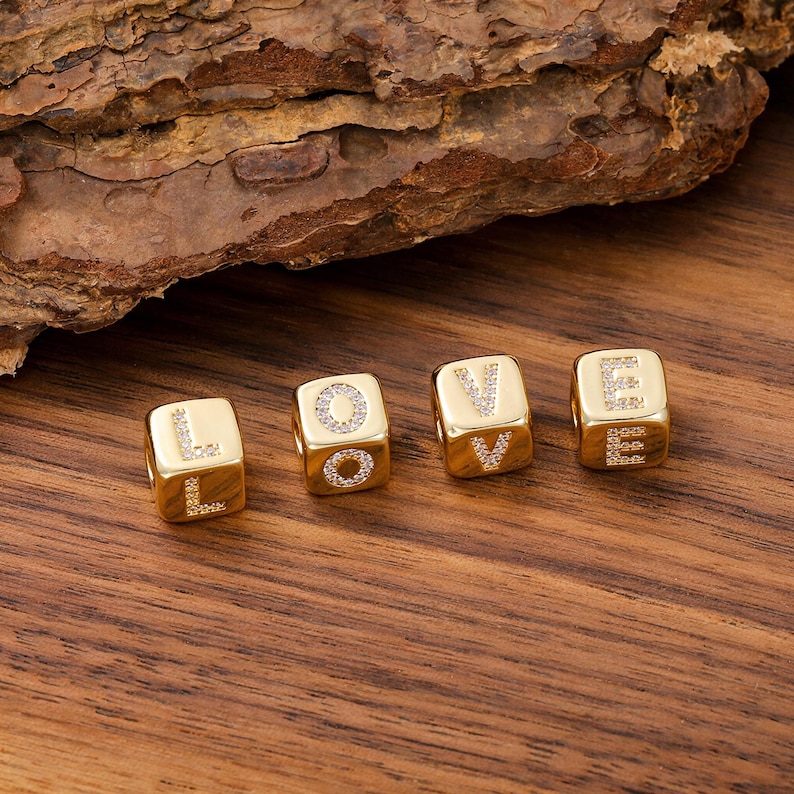 May include: Four gold-toned cube-shaped beads arranged to spell the word LOVE. Each bead features a letter formed by small, clear embedded stones on its front face. The beads have a smooth, polished metallic finish and a central hole for threading.