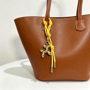 Puede incluir: Un bolso tote de cuero marrón con dos correas para el hombro y un accesorio de cuerda amarilla anudada sujeto a una correa. La cuerda sostiene un dije de perro globo de color dorado metálico con una superficie lisa y puntas de metal plateado.