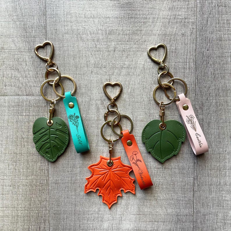 May include: Three keychains featuring heart-shaped clasps and rings. One has a dark green monstera leaf and a teal strap with floral engraving. Another has an orange maple leaf and an orange strap. The third has a green leaf and a pink strap.