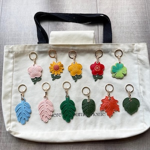May include: A collection of eleven leather-like keychains featuring various floral and leaf shapes in colors including pink, yellow, red, green, blue, and orange, each attached to a gold-tone metal ring, displayed on a cream canvas tote bag.