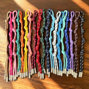 May include: A collection of fifteen colourful cord zip pulls, each featuring a series of decorative knots and finished with a metallic silver-toned cylindrical end cap. The cords are available in various solid colours including tan, pink, orange, yellow, red, green, blue, white, turquoise, purple, brown, and black.