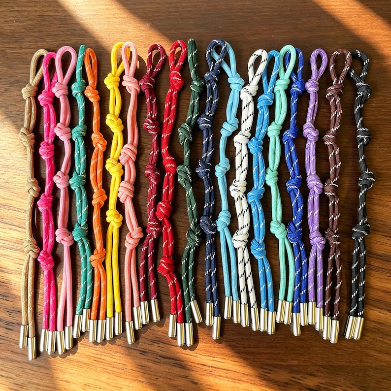 May include: A collection of fifteen colourful cord zip pulls, each featuring a series of decorative knots and finished with a metallic silver-toned cylindrical end cap. The cords are available in various solid colours including tan, pink, orange, yellow, red, green, blue, white, turquoise, purple, brown, and black.