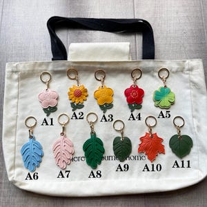 May include: Eleven leather-like keychains labeled A1 through A11, featuring various flower and leaf shapes in colors including pink, yellow, red, green, blue, and orange. Each keychain has a gold-tone metal ring and is displayed on a white tote bag.