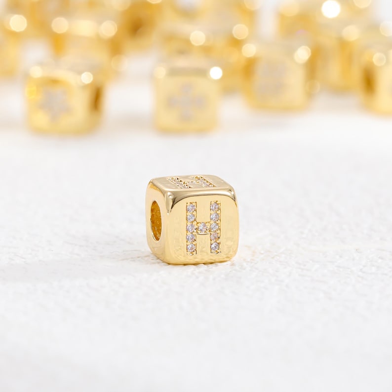 May include: A gold-toned cube-shaped bead featuring the letter H on its front face, embellished with small clear crystals. The bead has a smooth, polished metallic finish and a central hole for threading, with other similar beads blurred in the background.