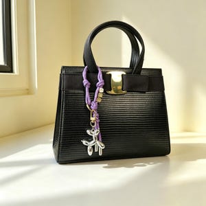 May include: A black structured handbag with a textured lizard-skin pattern, featuring two top handles, a horizontal black ribbon band, and a gold-tone metal clasp. A purple cord charm with small gold letter beads and a silver balloon dog pendant is attached.