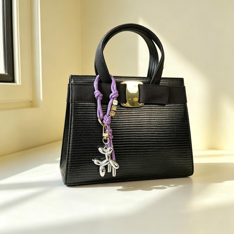 May include: A black structured handbag with a textured lizard-skin pattern, featuring two top handles, a horizontal black ribbon band, and a gold-tone metal clasp. A purple cord charm with small gold letter beads and a silver balloon dog pendant is attached.