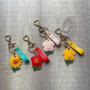May include: Four leather flower keychains with heart-shaped rings. Each features a unique flower shape in yellow, red, light pink, or yellow, paired with a matching leather strap embossed with a floral design and a name: Silk, Serene, Caitlin, or Serene.