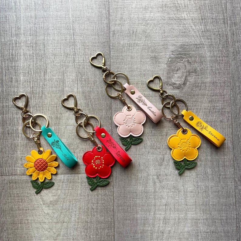 May include: Four leather flower keychains with heart-shaped rings. Each features a unique flower shape in yellow, red, light pink, or yellow, paired with a matching leather strap embossed with a floral design and a name: Silk, Serene, Caitlin, or Serene.