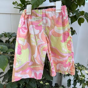 May include: Pink, yellow, and orange patterned baby pants with a button closure on the side. The pants are hanging on a white hanger. The tag reads "12-18m".