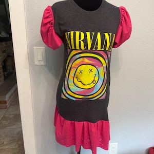 May include: A dark gray t-shirt dress featuring the yellow text NIRVANA above a graphic of a yellow smiley face with X eyes and a tongue sticking out, surrounded by a colorful swirl pattern. The dress has bright pink puff sleeves and a pink hem.