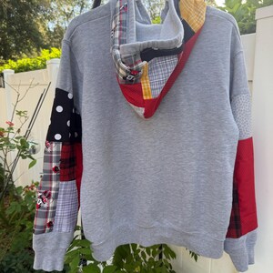May include: A grey long-sleeve hoodie featuring a patchwork design on the sleeves and hood. The patches include black and white polka dots, red and black plaid, grey plaid with Minnie Mouse graphics, yellow and white check, and solid red fabric.