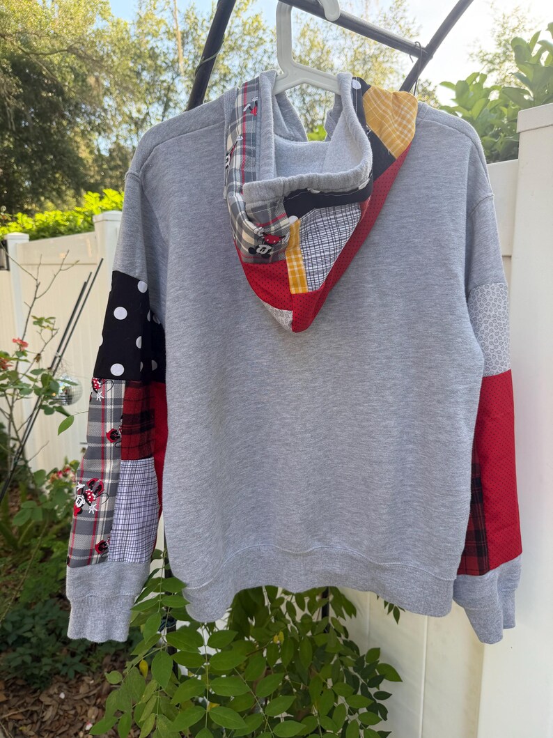 May include: A grey long-sleeve hoodie featuring a patchwork design on the sleeves and hood. The patches include black and white polka dots, red and black plaid, grey plaid with Minnie Mouse graphics, yellow and white check, and solid red fabric.