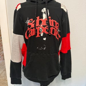 May include: A black hooded sweatshirt featuring a graphic print of a person and the red text Sabrina Carpenter. The garment is constructed with color-blocked panels in red, grey, and black, including leopard print accents on the shoulders.