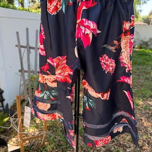 May include: Navy blue shorts with a vibrant floral pattern in shades of red, pink, and peach. The shorts have a sheer, striped detail at the hem. The garment is hanging on a white hanger.