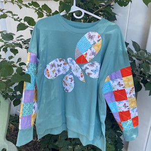 May include: A teal sweatshirt with a butterfly design made of patchwork fabric. The butterfly has various patterned wings, and the sleeves feature patchwork in squares of yellow, red, purple, and white. The sweatshirt is hanging on a white hanger.