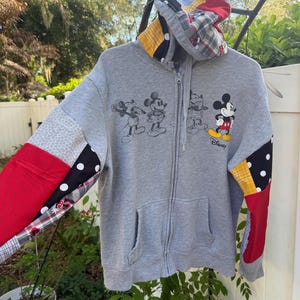 May include: A grey zip-up hoodie featuring a patchwork design on the sleeves and hood with red, yellow, black, and white patterns. The front displays a graphic of Mickey Mouse in various poses with the Disney logo printed in black ink.