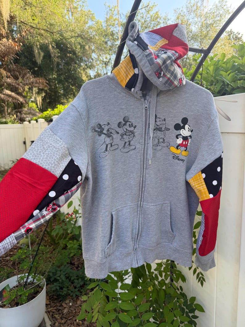May include: A grey zip-up hoodie featuring a patchwork design on the sleeves and hood with red, yellow, black, and white patterns. The front displays a graphic of Mickey Mouse in various poses with the Disney logo printed in black ink.