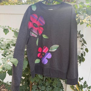 May include: Dark gray sweatshirt with appliqued floral design. The flowers are red, pink, and purple with green leaves. The sweatshirt is hanging on a black hanger against a white fence and greenery.