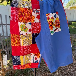 May include: Patchwork shorts with a vibrant mix of red, yellow, and blue paisley and floral patterns. One side features a patchwork design, while the other is solid blue with a floral pocket. The shorts are hanging on a white hanger.