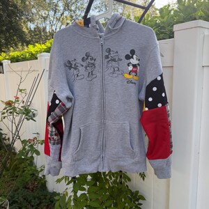 May include: A grey zip-up hoodie featuring a graphic of Mickey Mouse in various poses and the Disney logo. The sleeves are constructed with a patchwork design including black fabric with white polka dots, solid red panels, and grey plaid fabric.