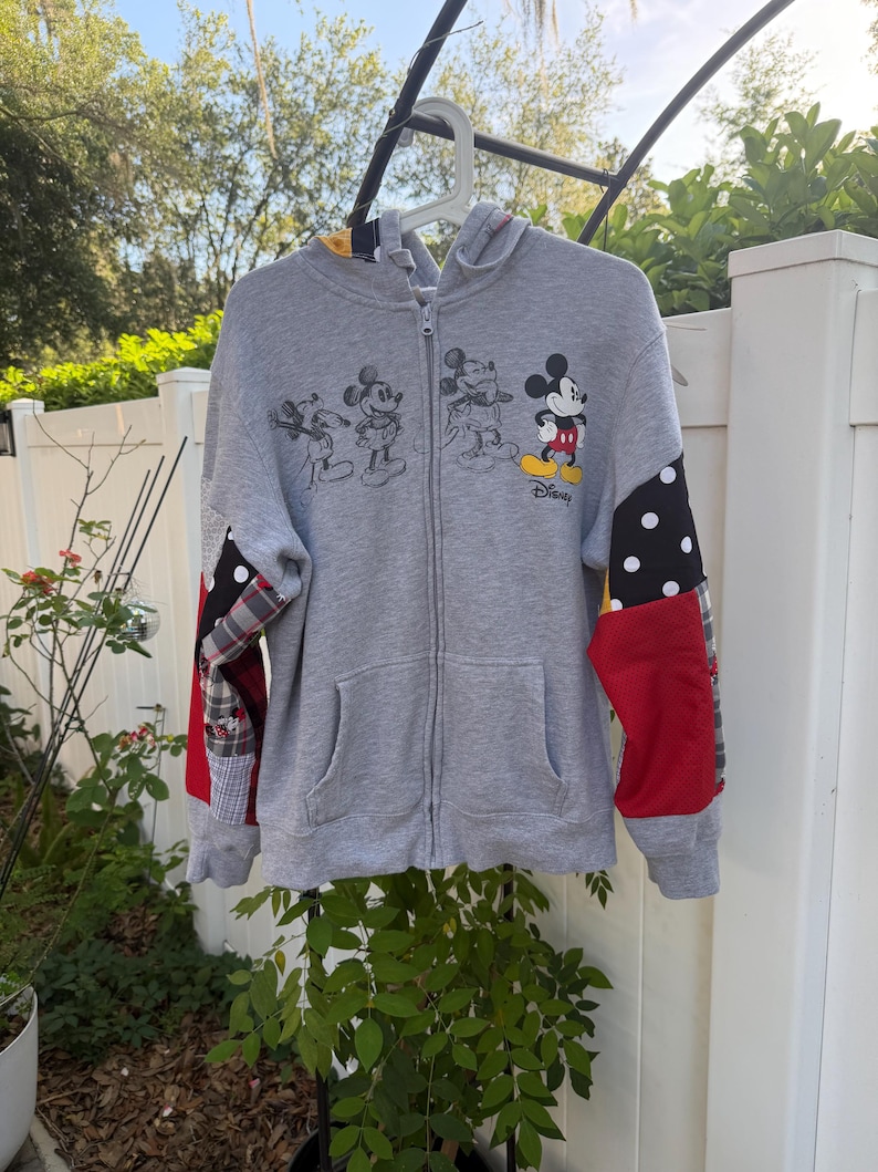 May include: A grey zip-up hoodie featuring a graphic of Mickey Mouse in various poses and the Disney logo. The sleeves are constructed with a patchwork design including black fabric with white polka dots, solid red panels, and grey plaid fabric.