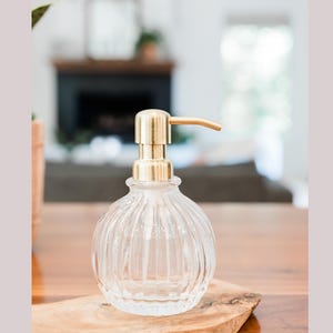 May include: Clear glass soap dispenser with a ribbed design and a gold-colored pump. The dispenser sits on a wooden surface, offering a touch of elegance to any bathroom or kitchen. The dispenser is approximately 6 inches tall.