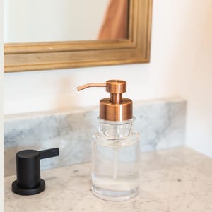 May include: Clear glass soap dispenser with a copper-colored pump and a black faucet. The dispenser is filled with a clear liquid and sits on a marble countertop. A gold-framed mirror is in the background.