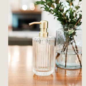 May include: A clear glass soap dispenser with vertical ridges and a gold-colored pump. The dispenser is on a wooden surface, with a blurred background featuring a fireplace and a vase with greenery.
