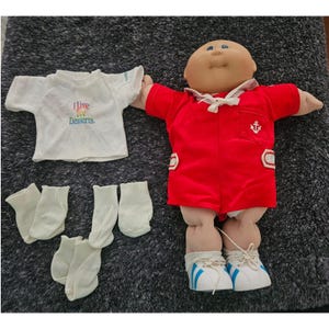 May include: A Cabbage Patch Kids doll with a round face, wearing a red sailor-style romper with a white anchor emblem, and white sneakers with blue stripes. Included are a white t-shirt with the text I Live For Desserts and three pairs of socks.