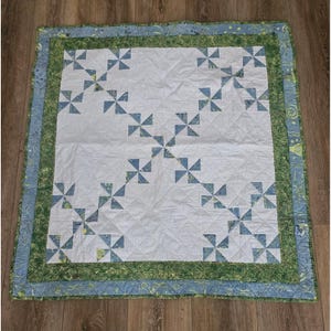 May include: A square quilt featuring a white center panel with a blue pinwheel pattern arranged in diagonal lines. The quilt is framed by a green border with a swirling line design and an outer light blue border with yellow and green accents.