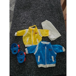 Puede incluir: Un conjunto de ropa para muñeca que incluye una chaqueta amarilla con cremallera, ribete blanco y un parche No.1, una chaqueta azul con cremallera, ribete blanco y un parche No.1, un pantalón blanco y patines de plástico azules con ruedas rojas.