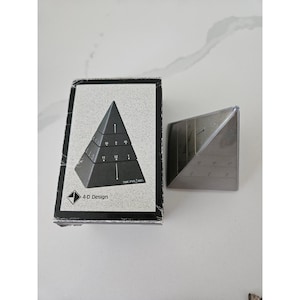 May include: A pyramid-shaped clock with a dark grey finish, featuring horizontal tiers marked with numbers and lines. The packaging box displays the product image, the text 4-D Design, and the text TIME PYRAMID, showing a tiered timekeeping display.