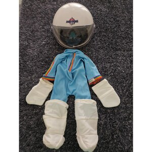 以下が含まれることがあります： 透明なバイザーと「Astronauts」の文字が入った白い宇宙飛行士のヘルメット。袖と中央に虹色のストライプのトリムがあしらわれた水色のジャンプスーツ、白い布製のミトン、白い布製のブーツがセットになった人形用のコスチューム一式。