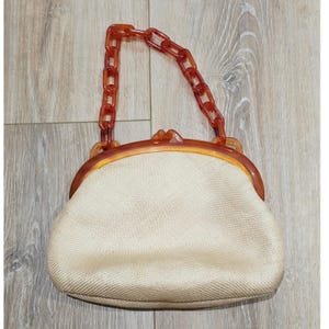 May include: A beige woven handbag featuring a semi-circular frame and a matching amber-colored resin chain handle. The bag has a kiss-lock closure mechanism integrated into the top frame, providing a structured shape with a textured fabric body.