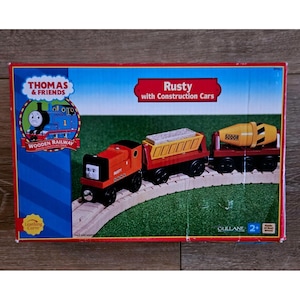 Może przedstawiać: Zapakowany drewniany zestaw kolejowy z Thomas & Friends. Zestaw zawiera czerwoną lokomotywę o nazwie Rusty, z wagonami budowlanymi. Na pudełku znajduje się napis "Rusty with Construction Cars" i "Wooden Railway". Zestaw przeznaczony jest dla dzieci w wieku 2+.