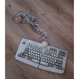 Puede incluir: Un teclado ergonómico dividido Lexmark de color beige con distribución QWERTY y una bola de seguimiento circular integrada en el centro. El dispositivo cuenta con un cable gris largo y enredado que sale de la parte superior con un conector estándar.