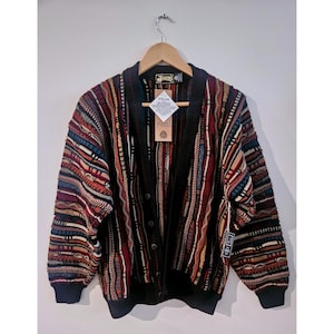 Puede incluir: Un cárdigan de manga larga con botones que presenta un complejo patrón de punto multicolor con rayas verticales en tonos rojo, azul, negro y beige. La prenda tiene un cuello, puños y dobladillo acanalados en color negro sólido.