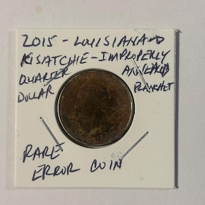 May include: A rare error coin, a 2015 Louisiana Kisatchie quarter dollar, improperly annealed planchet. The coin is displayed in a white card with handwritten text. The coin is a copper color.