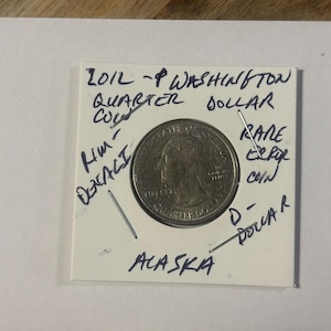 May include: A 2012-P Washington Quarter Dollar coin in a protective holder. The coin is silver-colored and features a profile of George Washington. Handwritten text on the holder indicates it is a rare coin from Alaska.